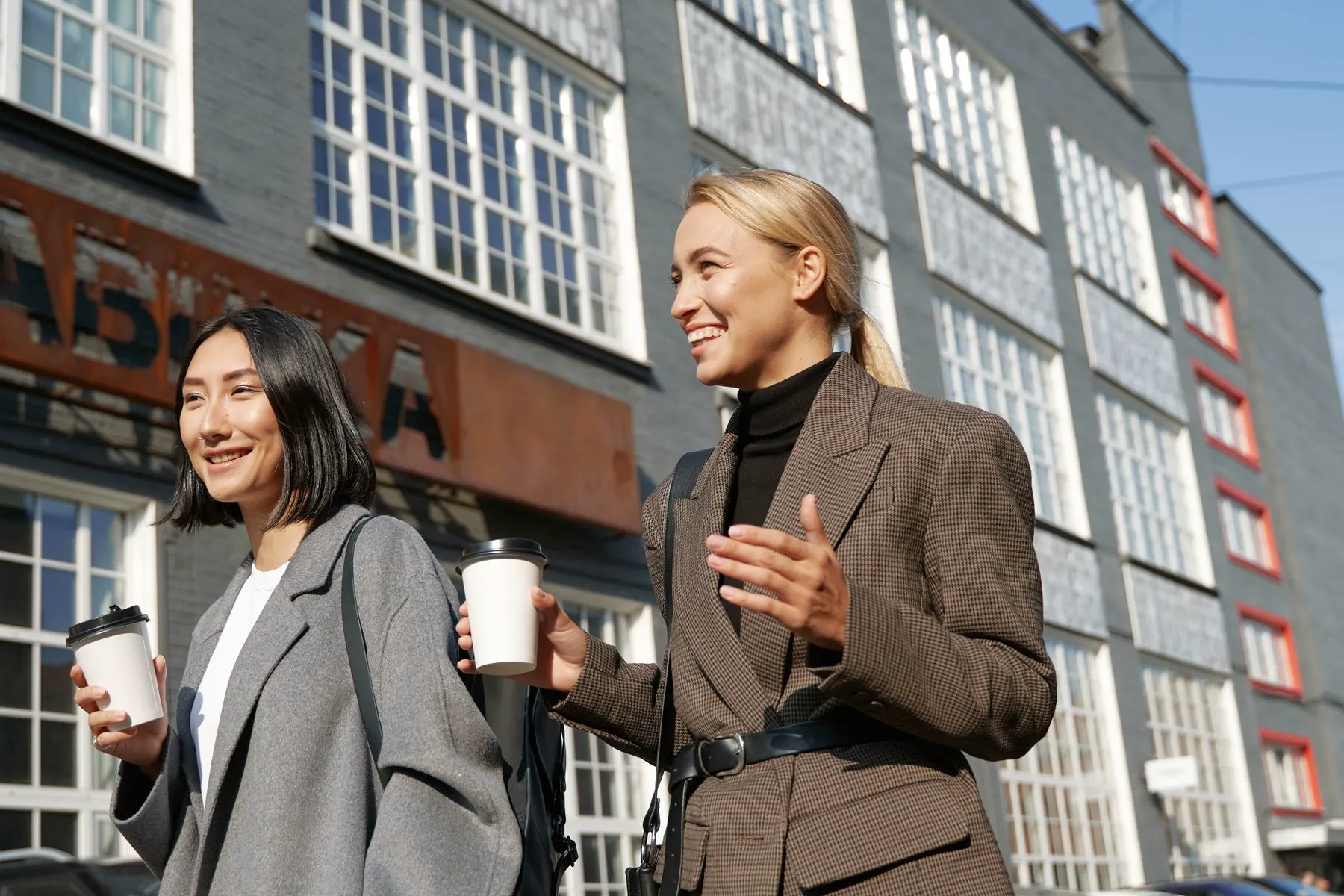 Twee vrouwen wandelen lachend met koffie over een zonnige straat